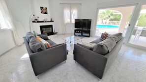 Uma sala de estar com sofá, mesa de centro de vidro, lareira, televisão e vista para uma piscina.