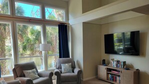 A living room with two armchairs, a blue ottoman, a bookshelf, and a TV mounted on the wall. - Egret’s Pointe Townhouse Sleeps 9 Amenities & POOLS included. GOLF CART (Edisto Island)