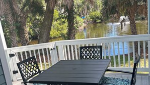 A wooden deck with a table and chairs, surrounded by lush greenery and a body of water. - Egret’s Pointe Townhouse Sleeps 9 Amenities & POOLS included. GOLF CART (Edisto Island)