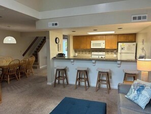 A compact kitchen with wooden cabinets, a microwave, and a refrigerator. There are three stools and a blue ottoman in the living area. - Egret’s Pointe Townhouse Sleeps 9 Amenities & POOLS included. GOLF CART (Edisto Island)