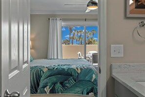 A bedroom with a bed, a view of palm trees, a bathroom with a marble countertop, and a ceiling light.