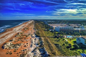 An aerial view of a coastal area with a beach, buildings, and a pool area.