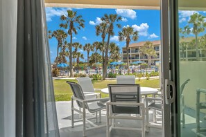 A balcony with white chairs and a table, overlooking a pool area with palm trees and umbrellas. - Luxury Beach Living In This Renovated Ground Floor Oceanfront 3BR Condo! (Saint Augustine)