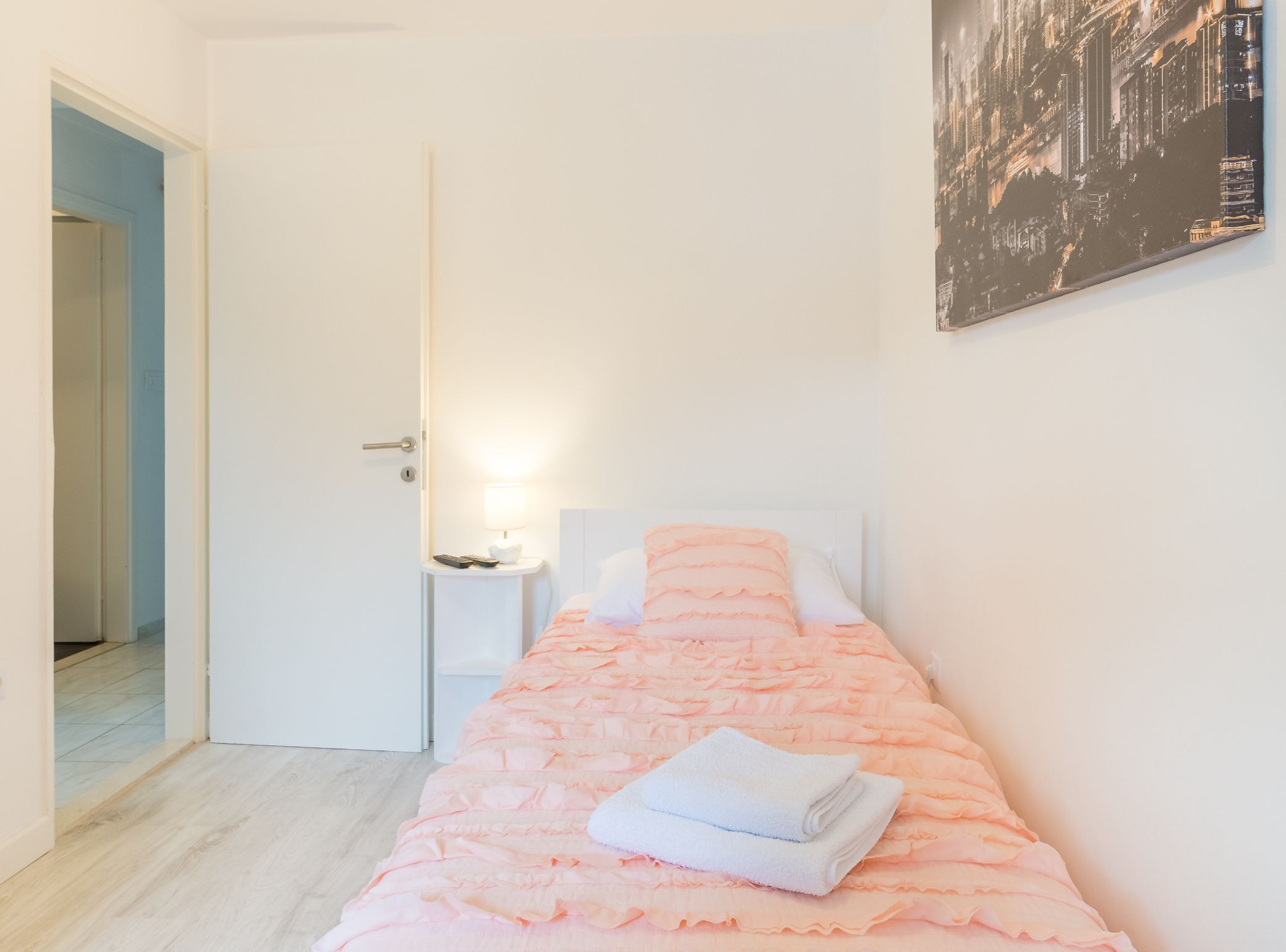 A neatly made bed with a pink bedspread and a white towel, a nightstand with a lamp and remote control, and a cityscape painting on the wall.