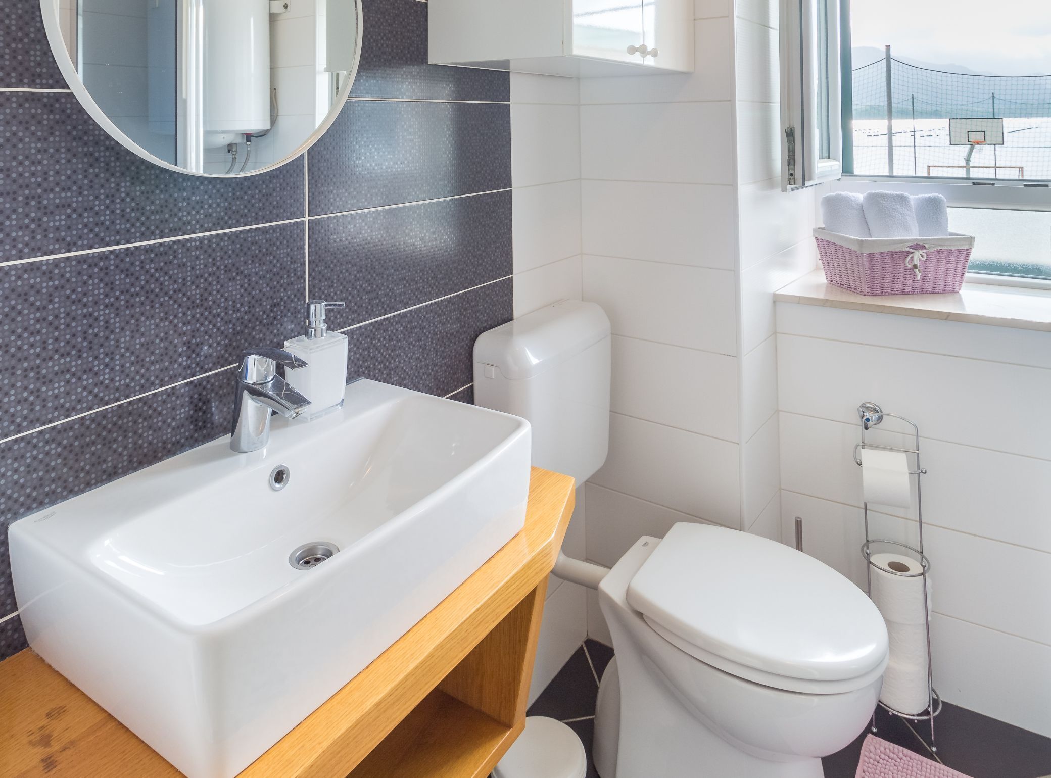 A bathroom with a white sink, a toilet, and a window with a view of a basketball court.