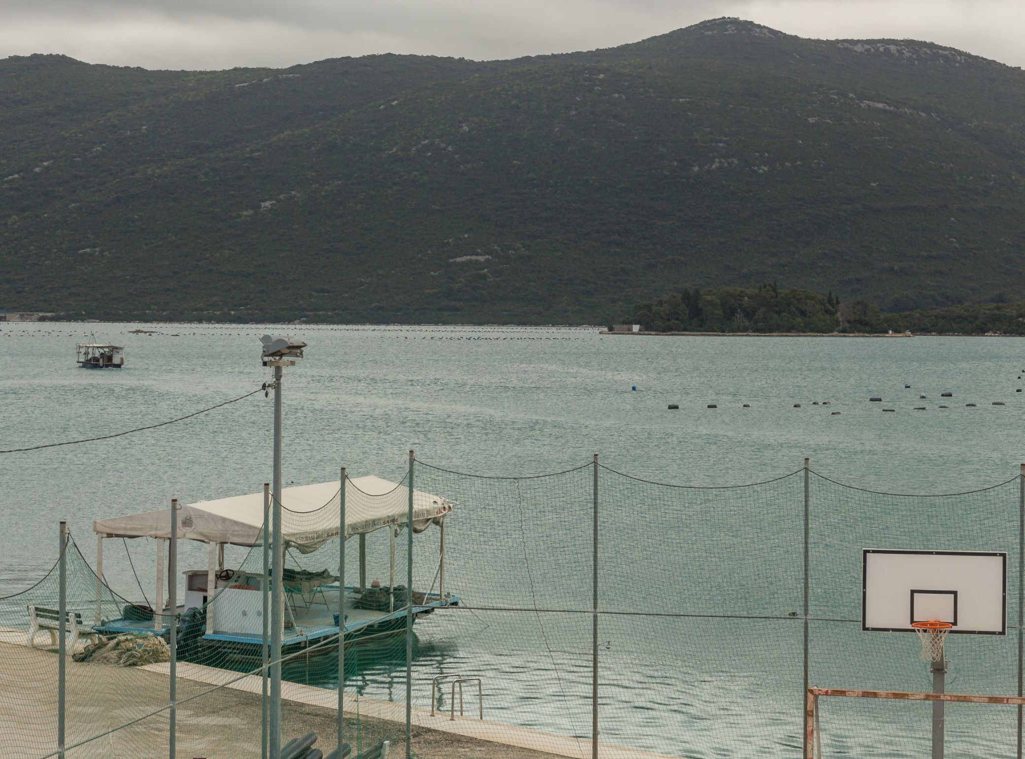 Un bateau était amarré près d’un terrain de basket-ball, entouré d’un filet, avec un panier et un paysage montagneux en arrière-plan.