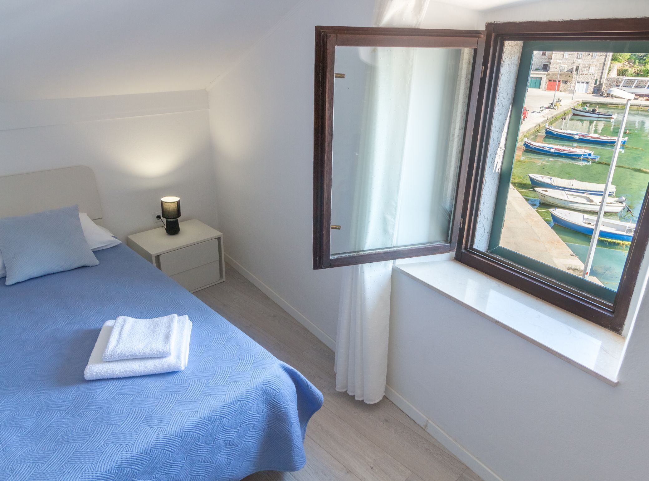 A bedroom with a bed, a nightstand, a lamp, and a window with a view of boats.