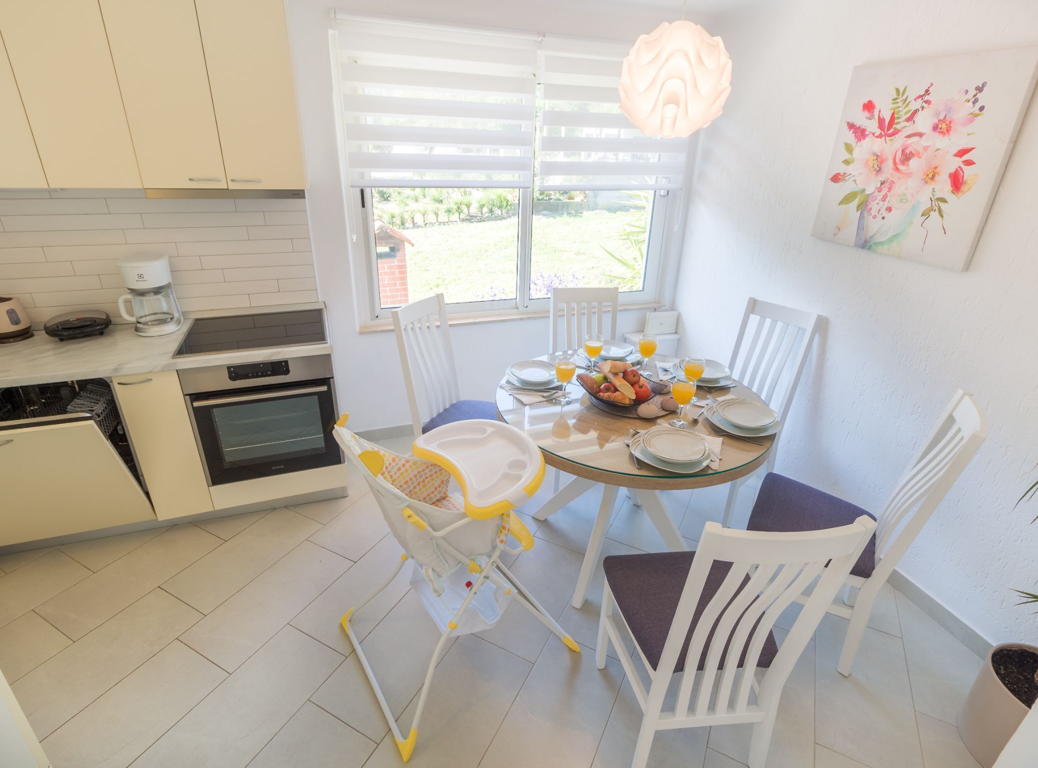 A bright kitchen with white cabinets, a dining table set for four, and a window with blinds.