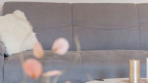 A grey sofa with a white fluffy pillow, a round white table with a candle and a remote control. - Holiday Home Riva (Ston)