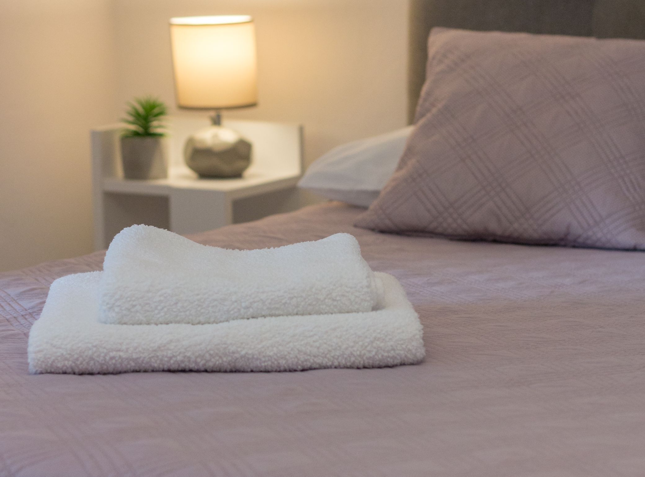 Una cama bien hecha con toallas blancas dobladas y una lámpara sobre la mesita de noche.