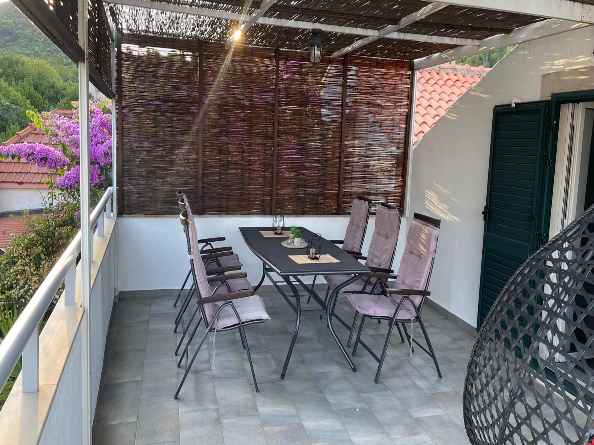 Ein Balkon mit Tisch und Stühlen, eine grüne Tür und Blick auf violette Blumen.