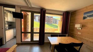 A room with a wooden table and chairs, a kitchen area, and a large sliding glass door leading to an outdoor area with a view of buildings and greenery. - 50m from ski lifts, 2nd floor, view ski slopes, balcony, tv, 28m², La Plagne (La Plagne)