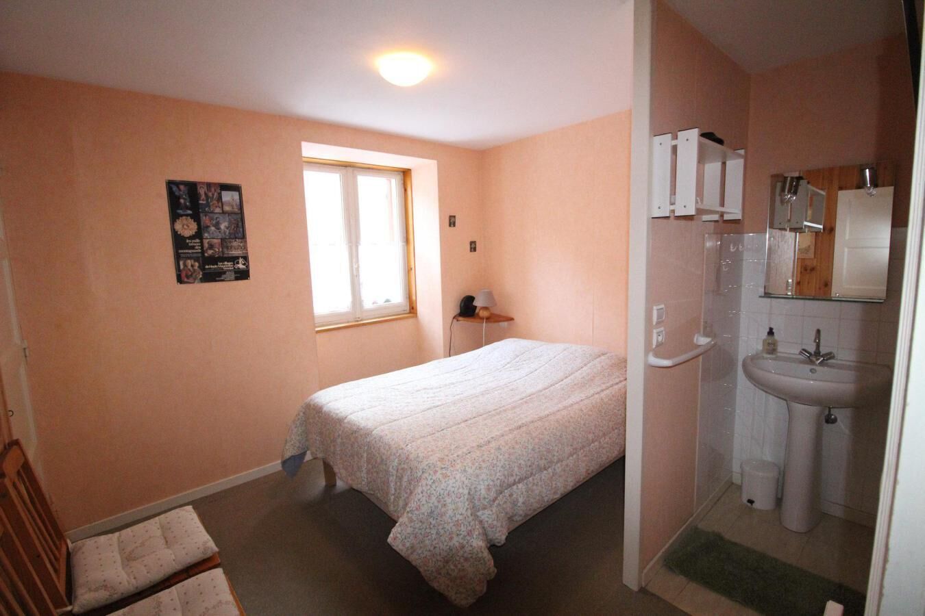 A bedroom with a bed, a window, a bedside table, and a bathroom with a sink and mirror.