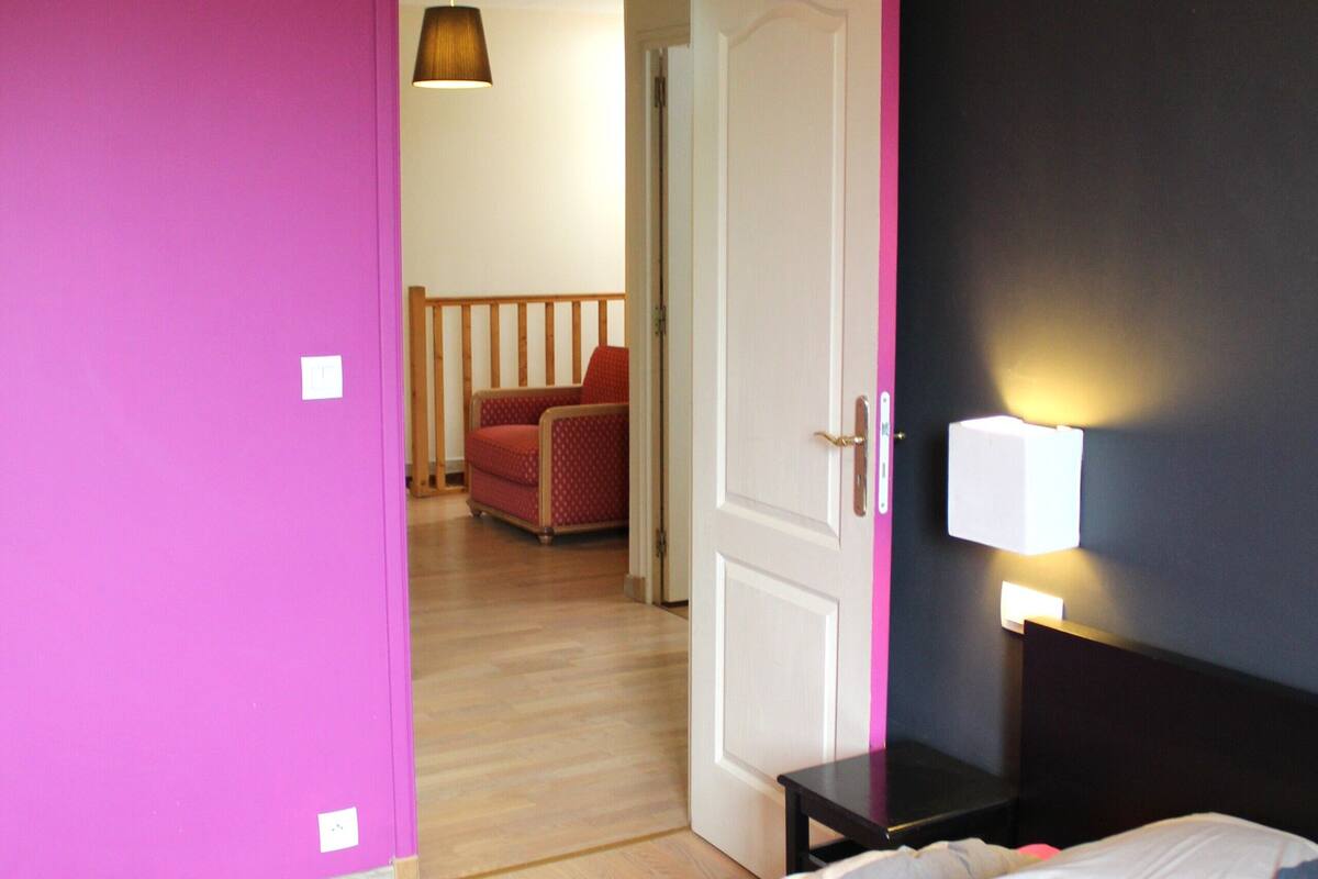 Une chambre à coucher comprenant un lit, une table de chevet, une lampe fixée au mur et une porte donnant sur une autre pièce.