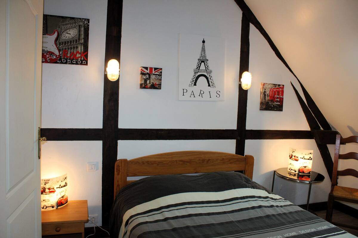 Une chambre à coucher comprenant un lit, une table de chevet, une lampe et des œuvres d’art murales, notamment des représentations de la tour Eiffel et du pont de Londres.