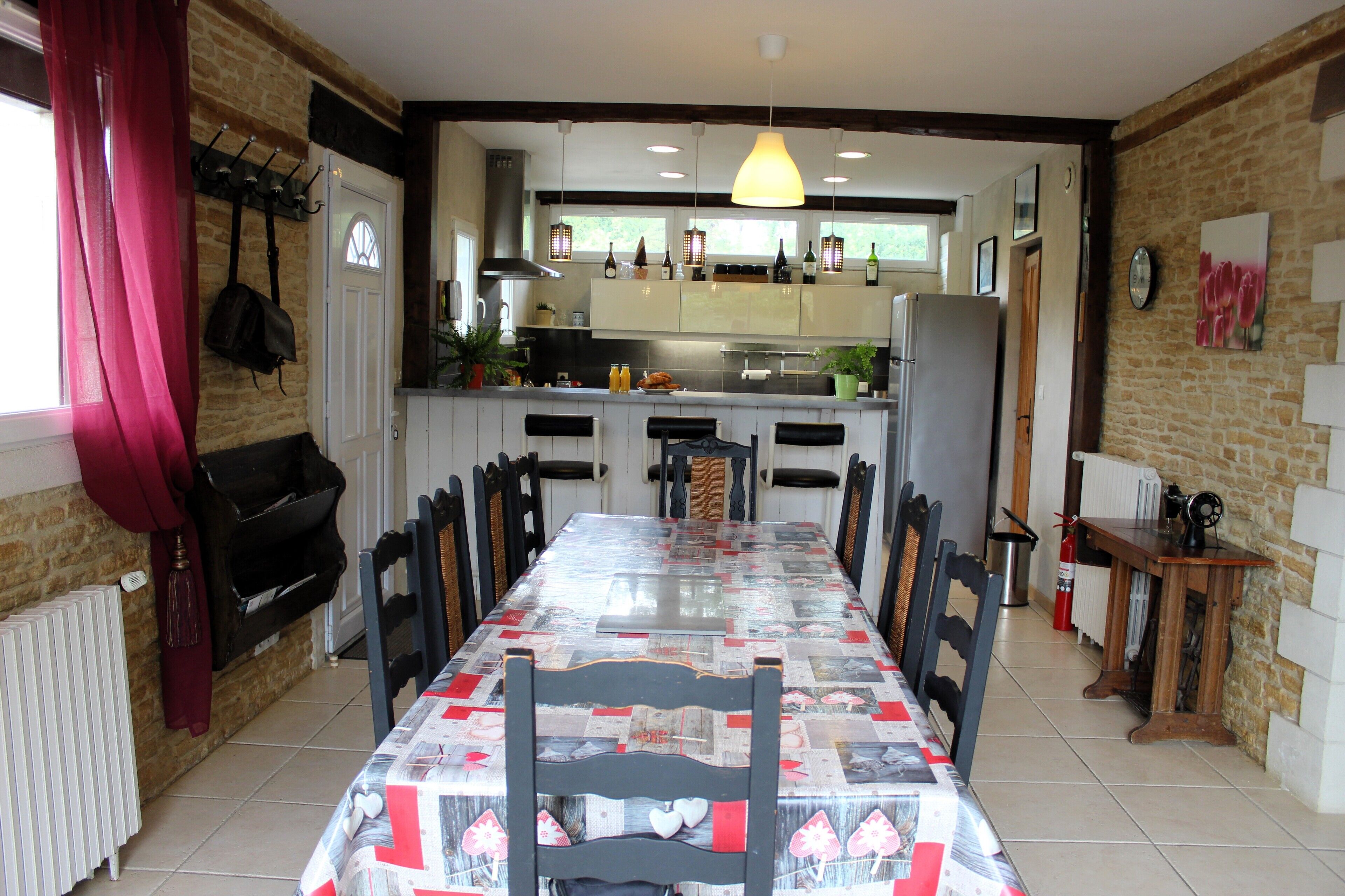 A dining area with a long table, chairs, a hanging light fixture, and a kitchen in the background.