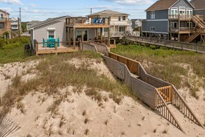Een huis aan het strand met een houten terras, een trap naar een balkon en uitzicht op het strand.