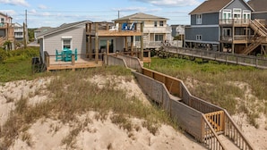 Een huis aan het strand met een houten terras, een trap naar een balkon en uitzicht op het strand.