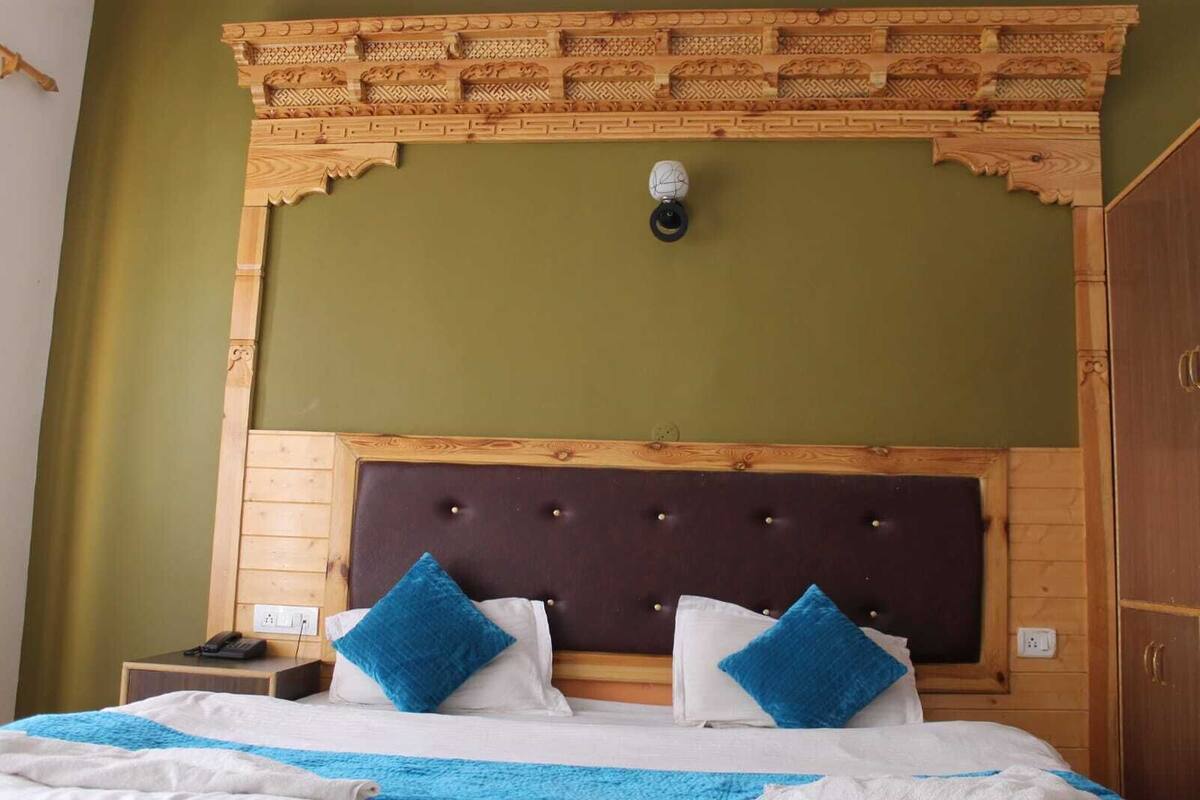 Habitación de hotel con cabecera de madera, cama con ropa de cama azul y blanca, y mesita de noche con un teléfono.