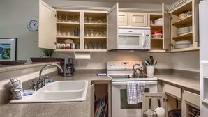 A compact kitchen with a sink, stove, microwave, and open cabinets containing dishes and utensils. - WW H202 Mather Street Retreat (NEW BRAUNFELS)
