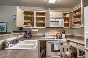 A compact kitchen with a sink, stove, microwave, and open cabinets containing dishes and utensils.
