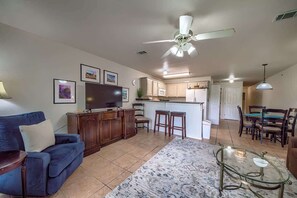 Uma sala de estar com sofá, televisão, ventilador de teto, cozinha com micro-ondas e área de jantar com cadeiras.