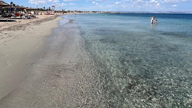 Een strand met helder ondiep water, parasols en ligbedden.