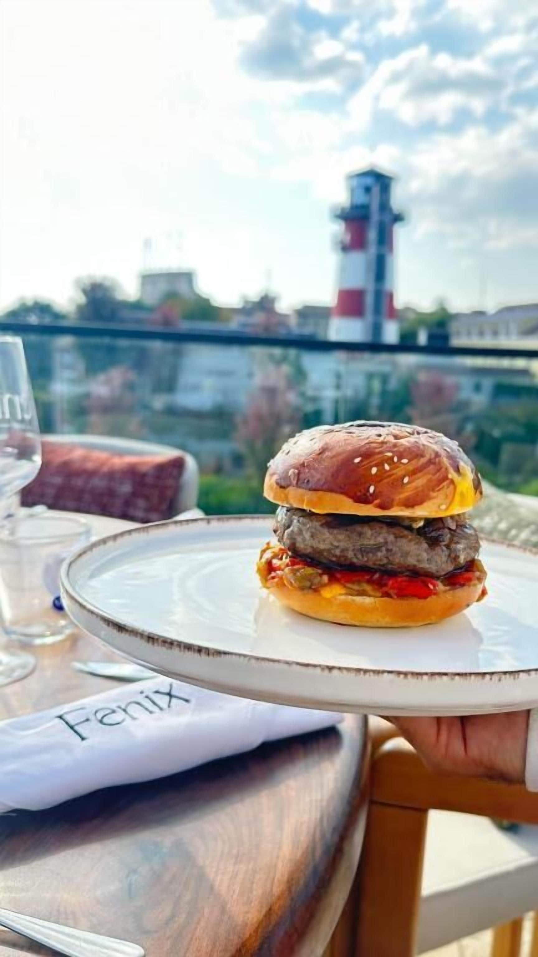 A burger on a white plate on a table with a view of a lighthouse in the background.
