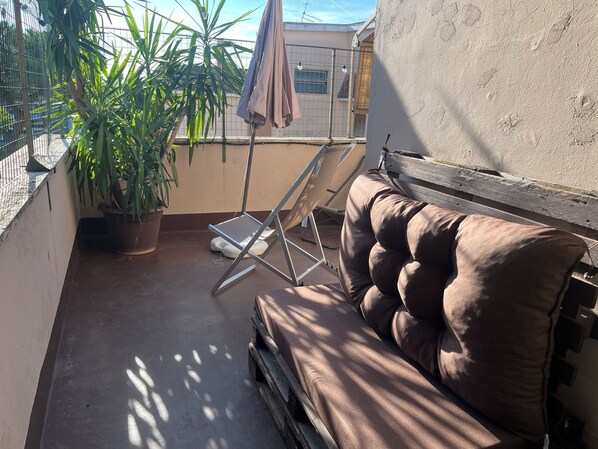 A small outdoor seating area with a cushioned bench and a metal folding chair. There is a potted plant and a metal fence in the background.