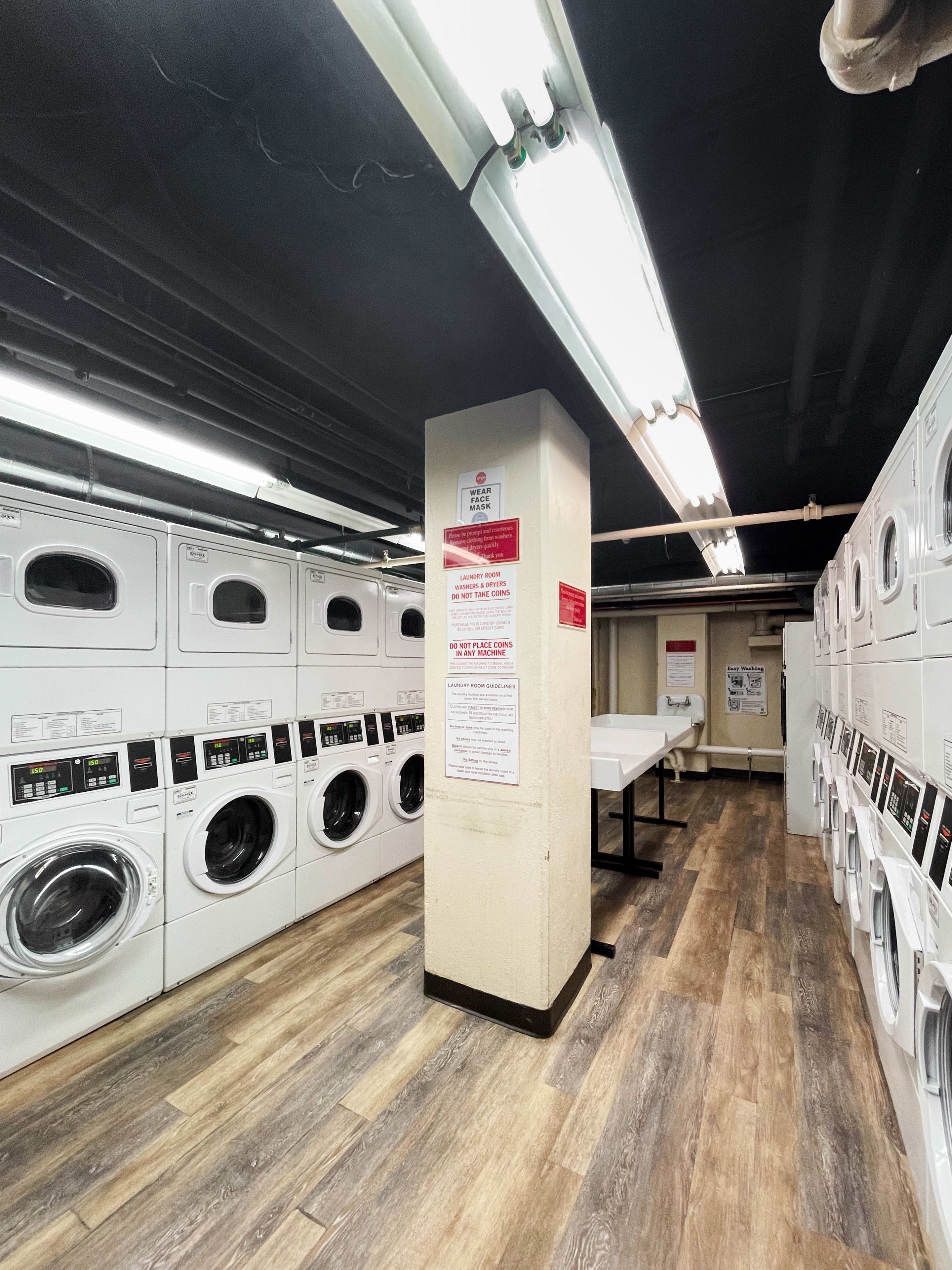 A laundry facility with multiple washing machines and dryers, wooden flooring, and signs indicating usage rules.