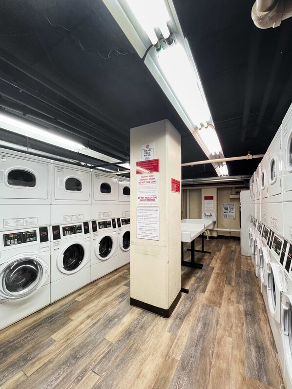 A laundry facility with multiple washing machines and dryers, wooden flooring, and signs indicating usage rules.
