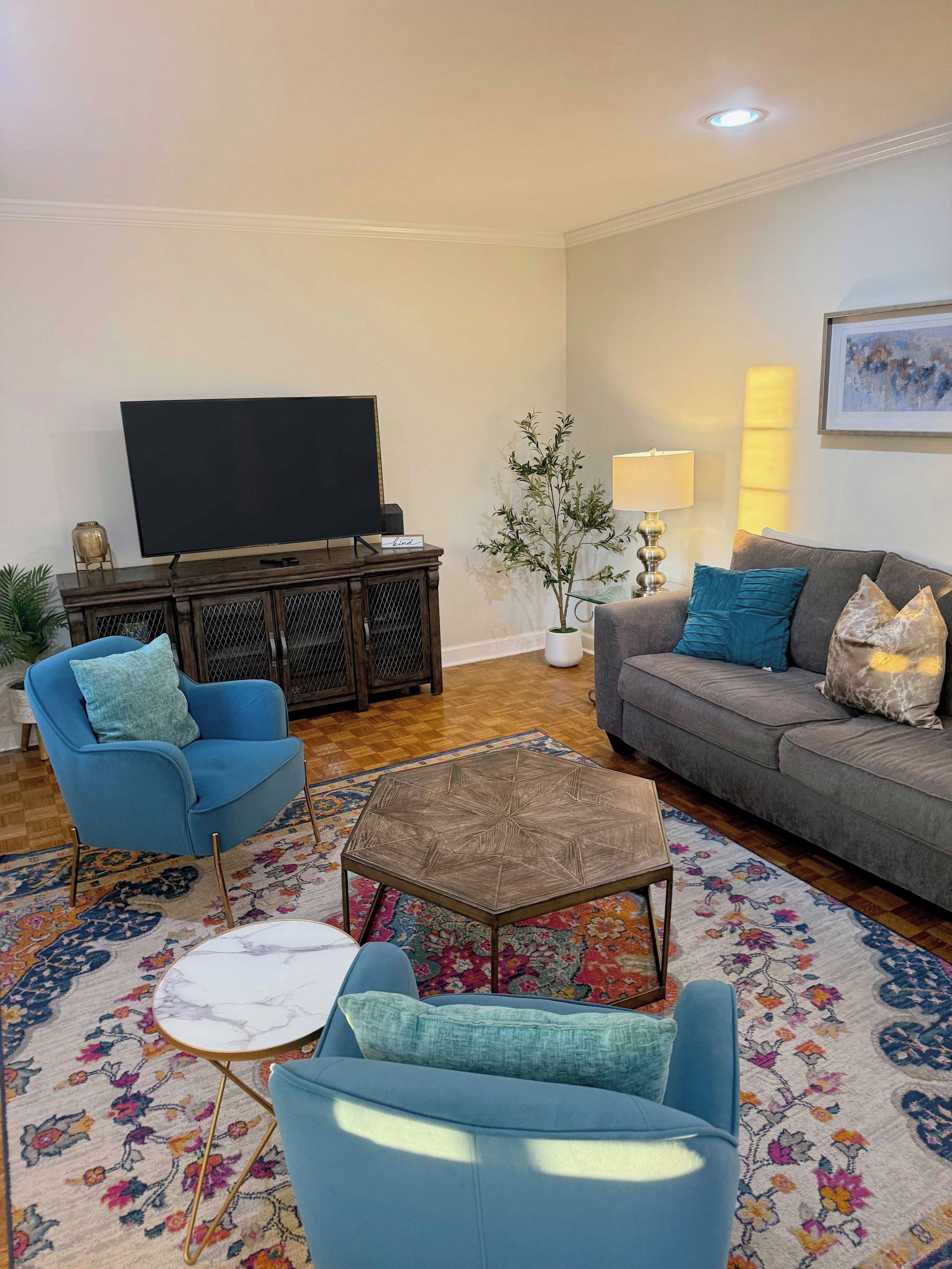 A living room with a flat-screen TV, a blue armchair, a grey sofa, a coffee table, and a patterned rug.