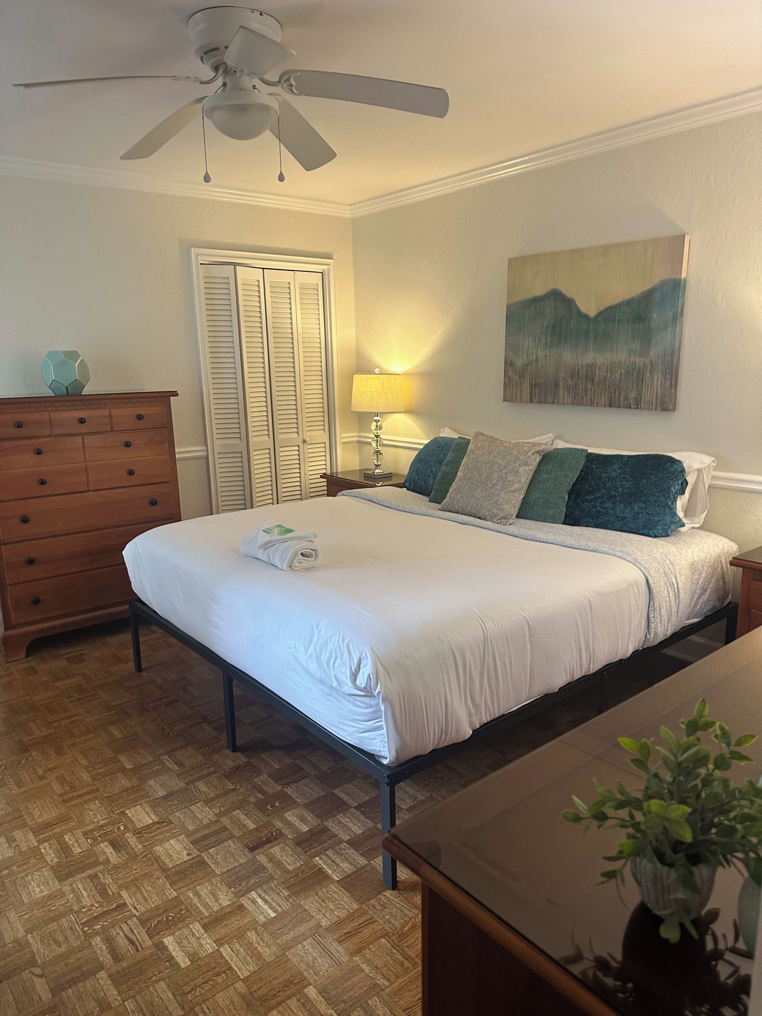 Ein Schlafzimmer mit Bett, Kommode, Nachttisch, Lampe und Deckenventilator.