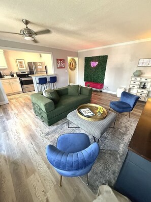 Una sala de estar moderna con un sofá verde, sillones azules, una mesa de centro y una cocina al fondo.
