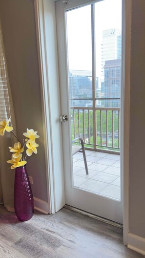A door leading to a balcony with a view of buildings and a vase with yellow flowers.