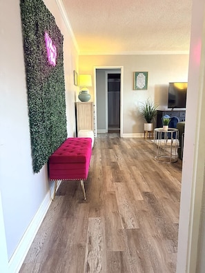 Una sala de estar con un pouf rojo, un adorno de pared verde, piso de madera, una lámpara, un televisor y una planta.