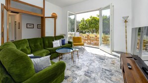 A modern living room with a green sofa, a round coffee table, and a wooden side table. There is a large glass door leading to an outdoor patio area with a dining table and chairs. - MGH Luxury - Villa Sirmione (Sirmione)