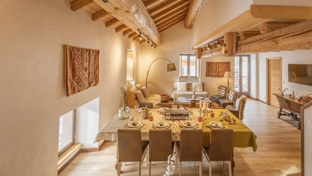 Un vaste espace dédié à la salle à manger, avec une longue table dressée pour le repas, des poutres en bois au plafond et de grandes fenêtres laissant entrer la lumière naturelle.