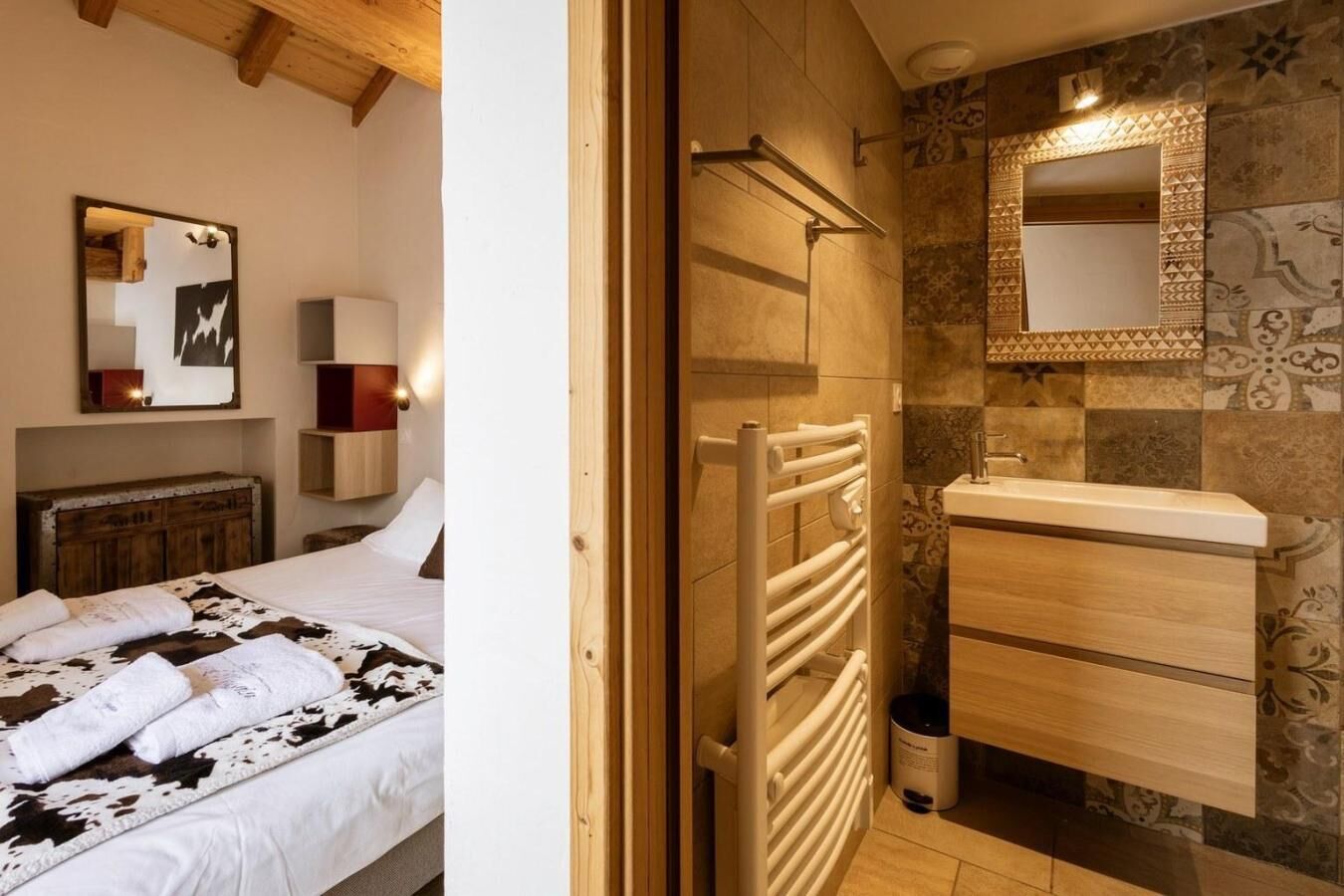 A bedroom with a bed, a bedside table, and a mirror. The bathroom has a sink, a mirror, and a towel rack.