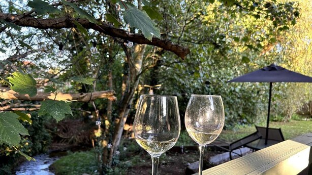 Two empty wine glasses on a wooden deck with a view of a stream and trees in the background. - Cosy and Secluded Brook-side Lodge (Sleaford)