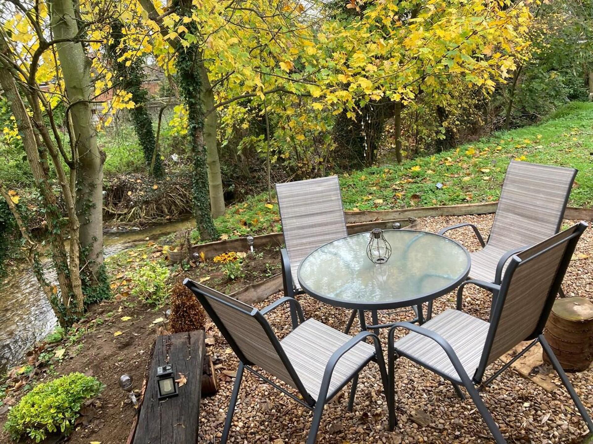 A round glass table with four chairs, surrounded by a garden with trees and a stream.