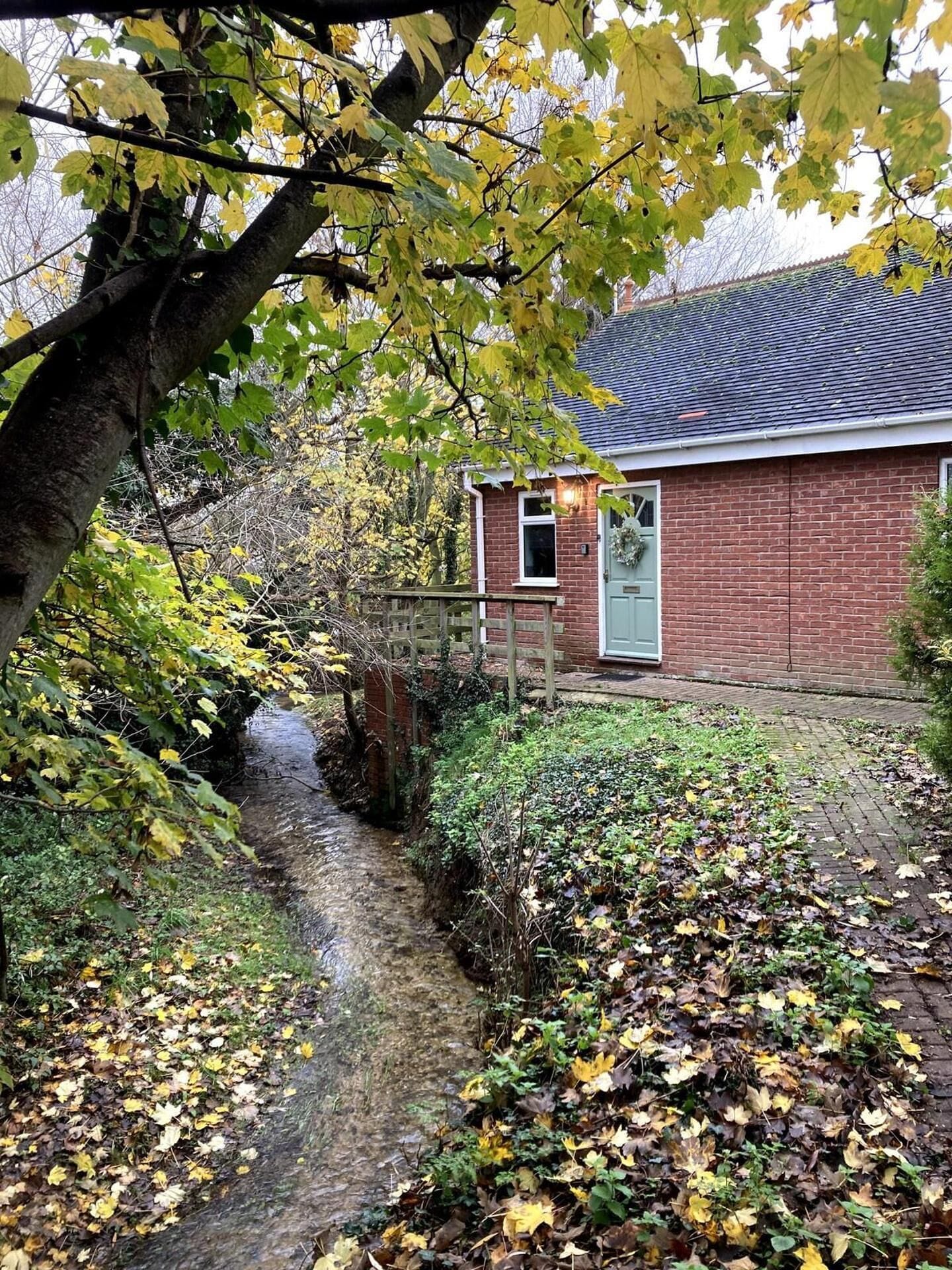 Une maison en briques avec un petit jardin devant, une clôture en bois et un ruisseau étroit.