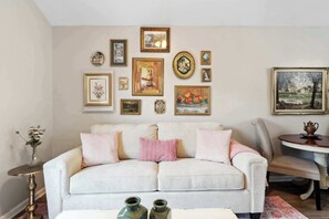 A living room with a light-colored sofa, a small round table with a teapot, and a wall decorated with various framed pictures. - Charming Home | Victorian District (Savannah)