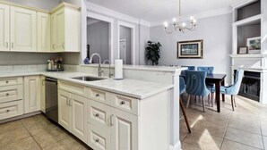 A modern kitchen with white cabinetry, a central island with a sink, and a dining area with blue chairs. - Charming Home, Victorian District (Savannah)