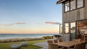 Una casa moderna con una terraza que da al océano, con un comedor de muebles de mimbre y una cocina visible a través de amplios ventanales.
