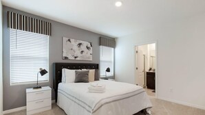 Une chambre à coucher comprenant un lit, des tables de chevet, une commode, une œuvre d’art accrochée au mur et une porte donnant accès à une salle de bain.