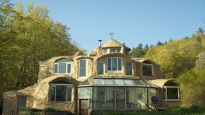 Una vivienda singular con forma de cúpula, varios ventanales y una sección cubierta de vidrio, rodeada de abundante vegetación.