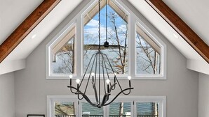 Une pièce avec un plafond haut, un lustre et de grandes fenêtres donnant sur des arbres et une étendue d’eau.