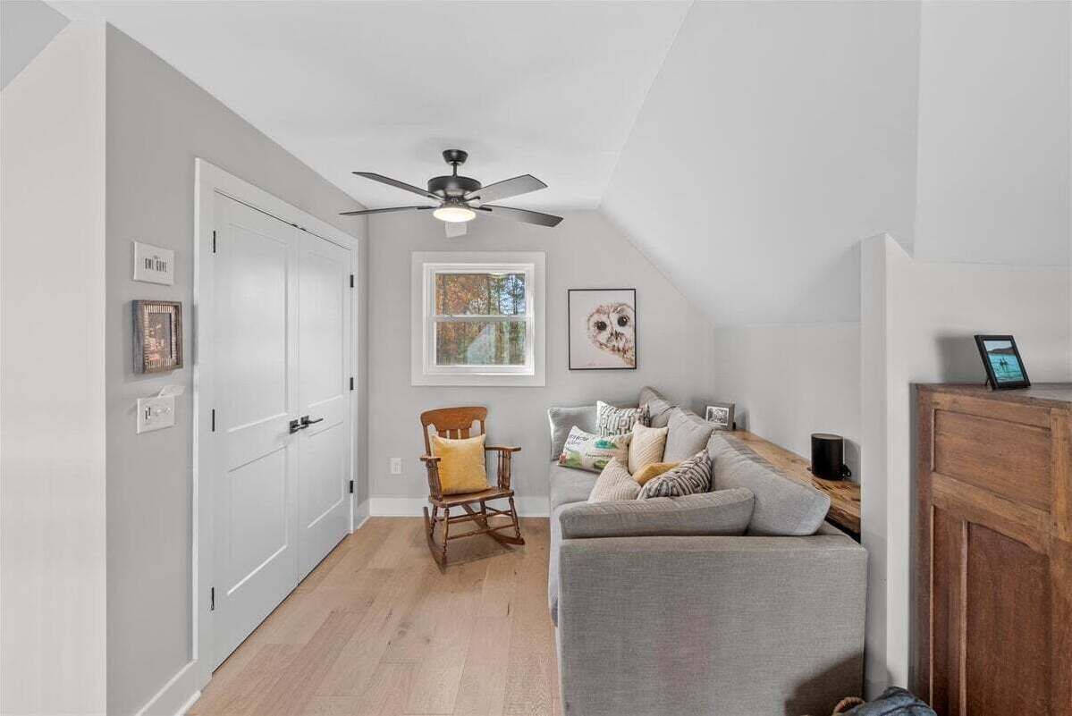 A room with a ceiling fan, a window, a grey sofa, a wooden rocking chair, and a white door.