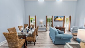 Un coin repas avec des chaises en osier et une table en verre, un canapé bleu et un tableau représentant des palmiers.